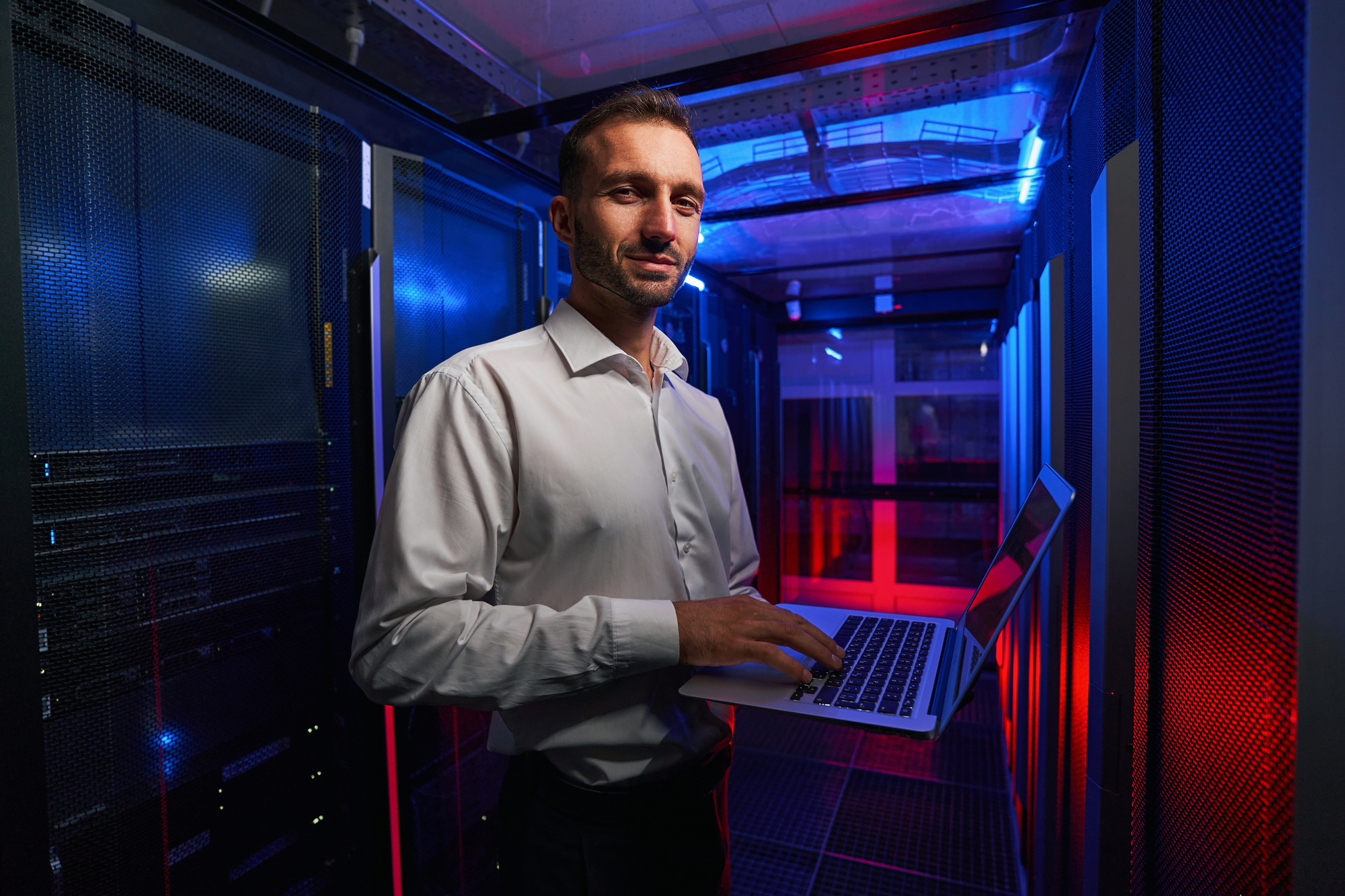 Bearded smiling system administrator is working with server rack and telecommunication equipments in